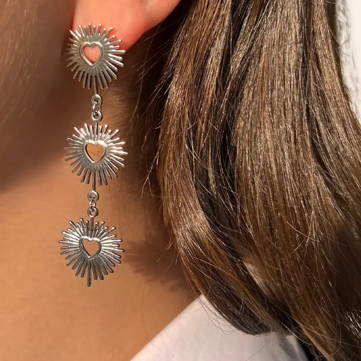 Boucles d'oreilles Dubaï en acier inoxydable argenté portées, design dynamique avec pendentifs solaires et cœurs ajourés, capturant l'opulence et le glamour de Dubaï.
