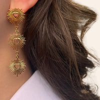 Boucles d'oreilles Dubaï dorées portées, pendentifs solaires et cœurs ajourés capturant la lumière et l'énergie de Dubaï, ajoutant une touche glamour à votre look.